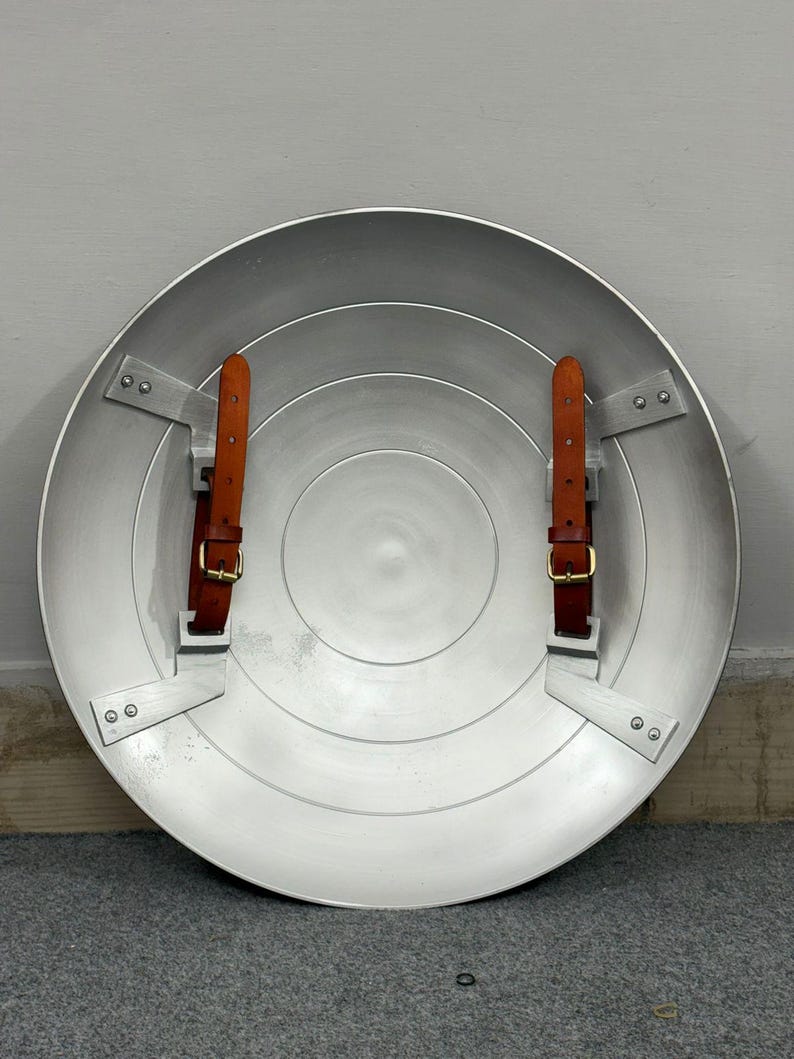 Puede incluir: Un escudo circular de metal plateado con un dise&ntilde;o liso de anillos conc&eacute;ntricos. El interior incluye dos correas de cuero marr&oacute;n con hebillas de metal dorado, cada una asegurada al escudo mediante soportes y remaches de metal plateado.