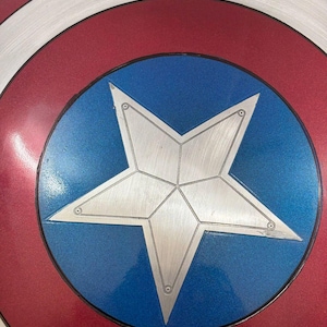 May include: A circular shield featuring a central silver star on a blue background, surrounded by a white ring and an outer red ring. The surface has a metallic finish with visible concentric circular textures and small rivets on the star points.