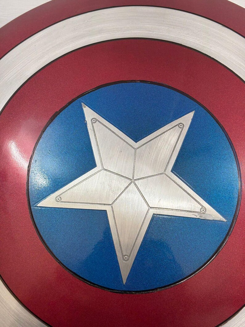 May include: A circular shield featuring a central silver star on a blue background, surrounded by a white ring and an outer red ring. The surface has a metallic finish with visible concentric circular textures and small rivets on the star points.