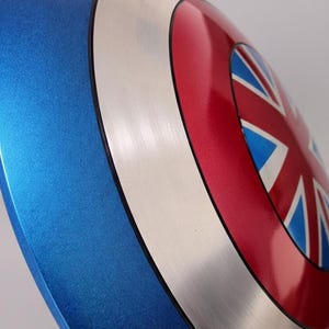 Op de afbeelding: Een metallic blauw, zilver en rood schild met een Union Jack-ontwerp. Het schild heeft een gebogen vorm en een glanzende afwerking. Het schild is een replica van de Britse vlag.
