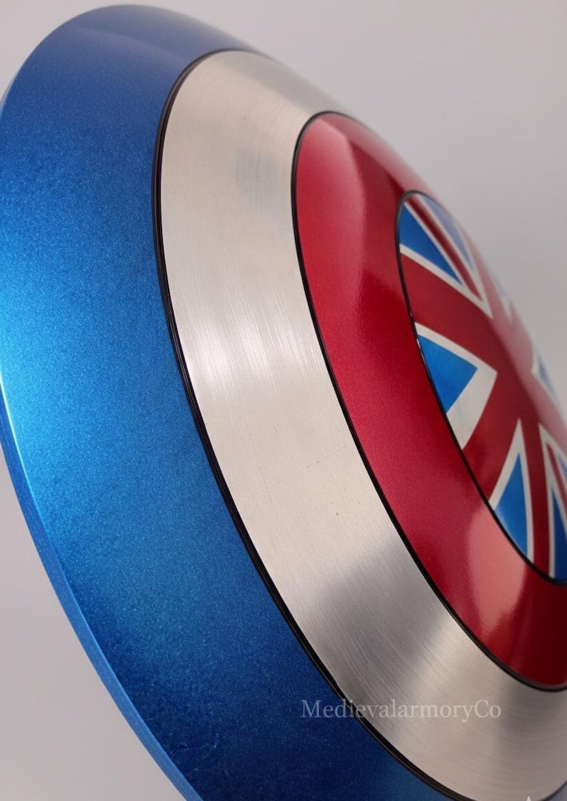 Op de afbeelding: Een metallic blauw, zilver en rood schild met een Union Jack-ontwerp. Het schild heeft een gebogen vorm en een glanzende afwerking. Het schild is een replica van de Britse vlag.