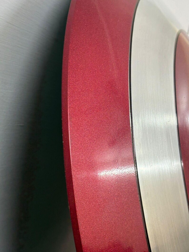 May include: A circular shield featuring alternating concentric rings of metallic red and brushed silver. The surface displays a smooth, reflective finish with a distinct color contrast between the deep red sections and the bright silver bands.