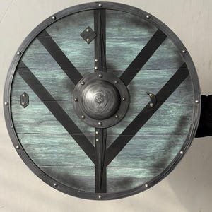 May include: A circular wooden shield with a teal and grey finish, featuring black metal accents and a central boss. The shield has a metal rim and is adorned with rivets. The design includes diagonal black stripes.