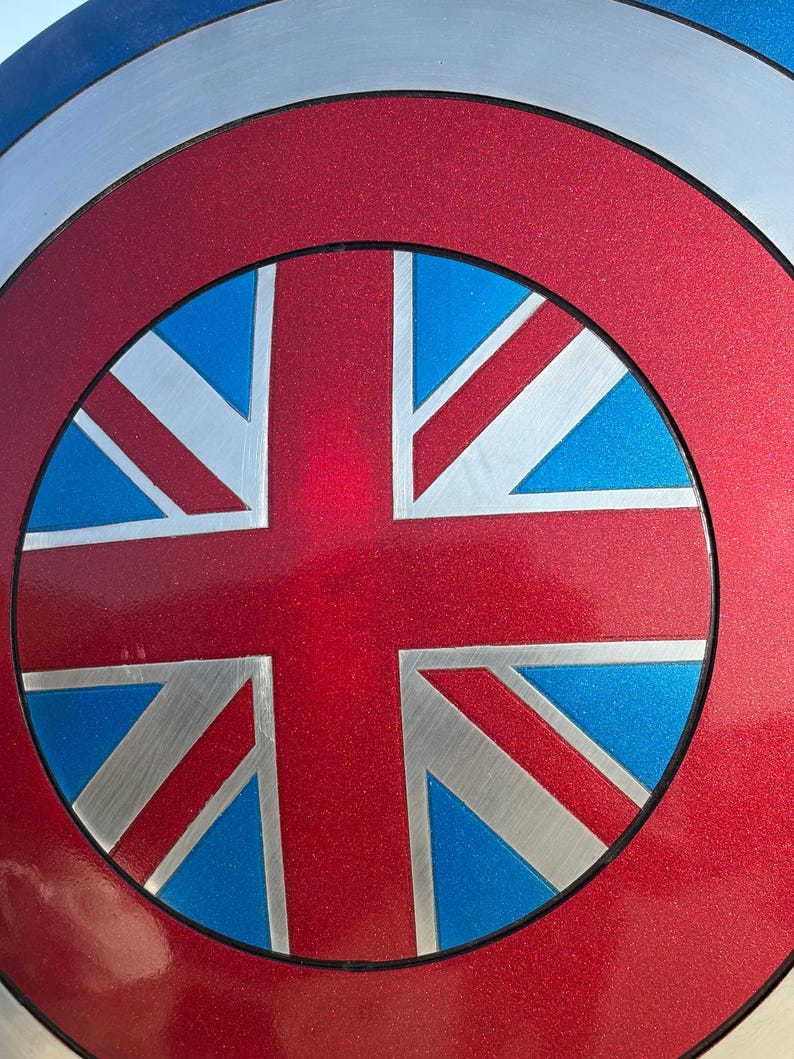 Op de afbeelding: Een metalen schild met een rood, wit en blauw ontwerp. Het midden toont een rode cirkel met een Union Jack vlagontwerp in zilver, blauw en rood. De buitenste ring is zilver, wat een patriottisch, superheldenachtig effect cre&euml;ert.