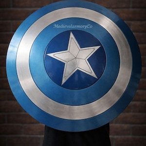 Op de afbeelding: Een rond schild met een blauw en zilver ontwerp. Het schild heeft een grote witte ster in het midden, omringd door concentrische cirkels van blauw en zilver. Het schild wordt voor een persoon gehouden, met een bakstenen muur op de achtergrond.