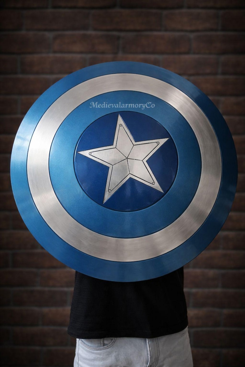 Op de afbeelding: Een rond schild met een blauw en zilver ontwerp. Het schild heeft een grote witte ster in het midden, omringd door concentrische cirkels van blauw en zilver. Het schild wordt voor een persoon gehouden, met een bakstenen muur op de achtergrond.