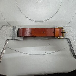 May include: A brown leather strap with a brass-toned metal buckle attached to two silver-colored, textured metal brackets. The strap features a smooth finish with visible stitching and a single prong buckle mechanism for securing the leather band.