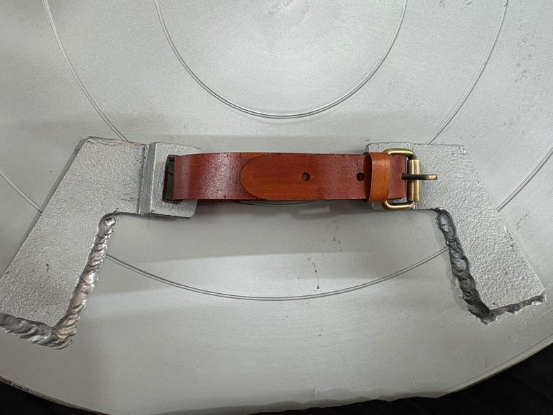 May include: A brown leather strap with a brass-toned metal buckle attached to two silver-colored, textured metal brackets. The strap features a smooth finish with visible stitching and a single prong buckle mechanism for securing the leather band.