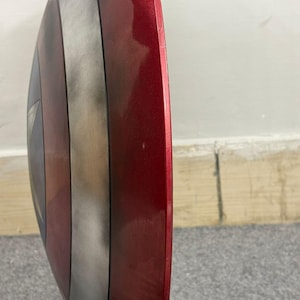 Puede incluir: Una vista de perfil de un escudo circular que presenta un borde exterior rojo met&aacute;lico, un anillo central plateado y una secci&oacute;n central azul oscuro. La superficie tiene un acabado liso y reflectante con una ligera curvatura.