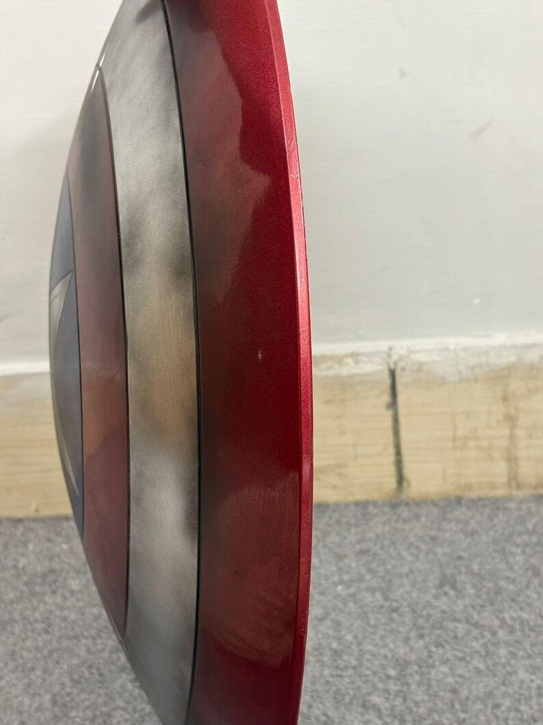 Puede incluir: Una vista de perfil de un escudo circular que presenta un borde exterior rojo met&aacute;lico, un anillo central plateado y una secci&oacute;n central azul oscuro. La superficie tiene un acabado liso y reflectante con una ligera curvatura.