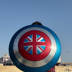 Op de afbeelding: Een rond schild met een blauw, zilver en rood kleurenschema. Het midden heeft een rode cirkel met een Union Jack-ontwerp. De buitenrand is blauw en het schild heeft een metallic uitstraling.