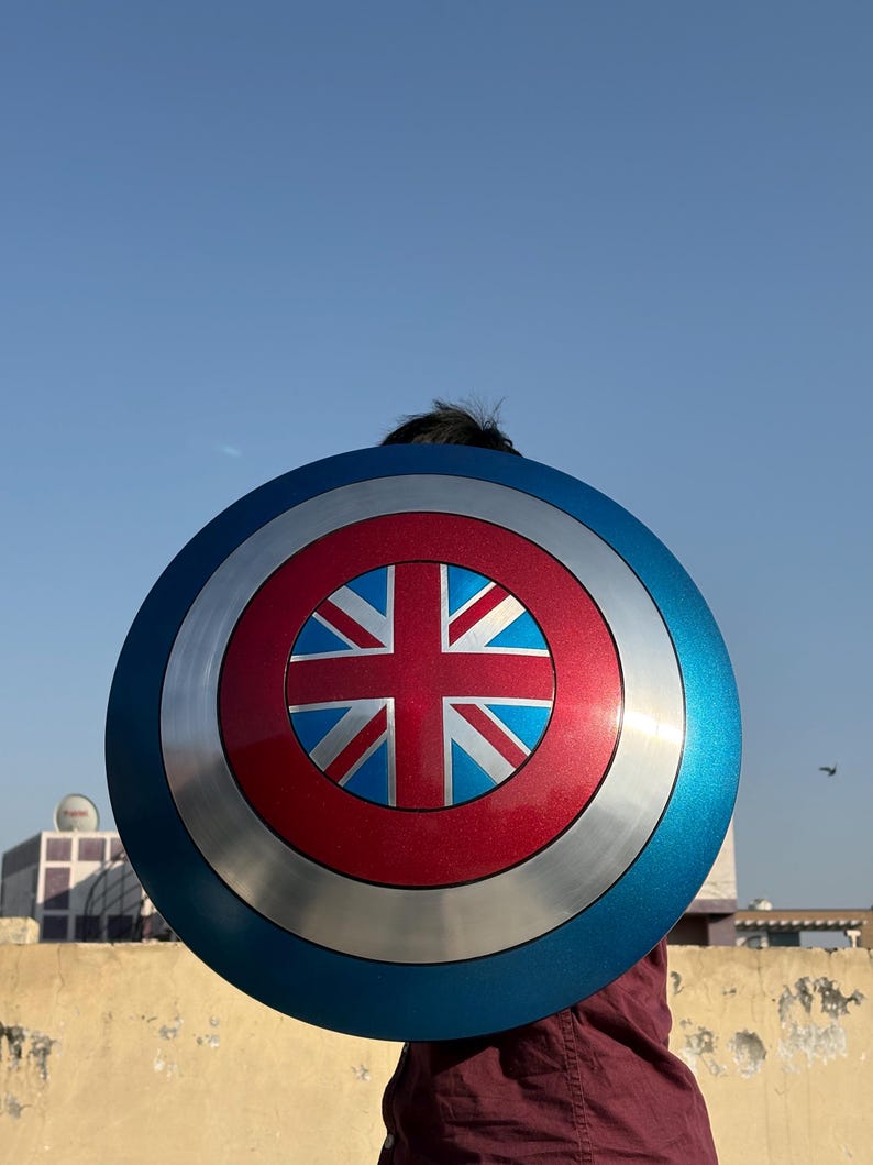 Op de afbeelding: Een rond schild met een blauw, zilver en rood kleurenschema. Het midden heeft een rode cirkel met een Union Jack-ontwerp. De buitenrand is blauw en het schild heeft een metallic uitstraling.