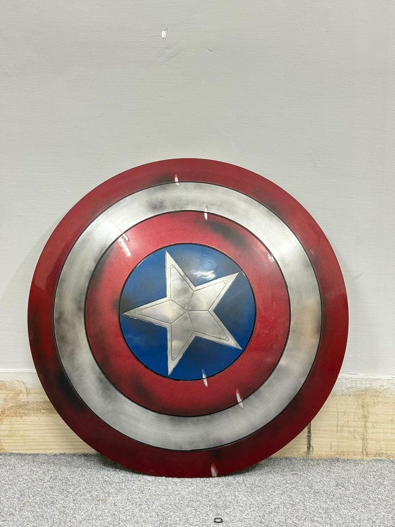 Puede incluir: Un escudo circular con una estrella plateada central sobre un fondo azul, rodeado por un anillo plateado y un anillo exterior rojo. La superficie tiene un acabado desgastado con manchas oscuras y ara&ntilde;azos sobre la textura met&aacute;lica.