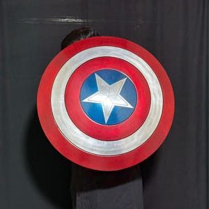 May include: A round shield with a red, white, and blue color scheme. The shield features a central blue circle with a white star, surrounded by concentric silver and red bands. The shield is a prop or costume accessory.