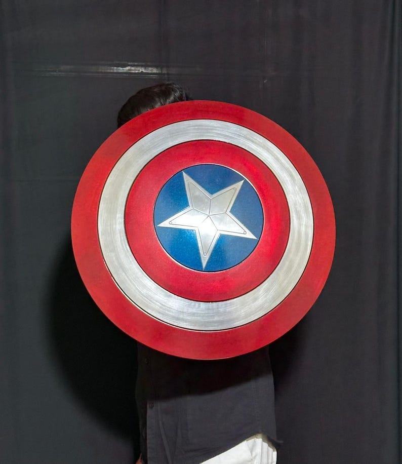 May include: A round shield with a red, white, and blue color scheme. The shield features a central blue circle with a white star, surrounded by concentric silver and red bands. The shield is a prop or costume accessory.