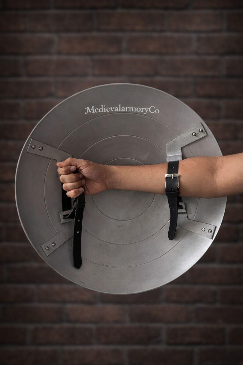 Op de afbeelding: Een zilverkleurig, rond schild met een geklonken metalen handvat en armriem. Het schild heeft een licht verweerde uitstraling en de tekst "MedievalarmoryCo" is zichtbaar. De achtergrond is een bakstenen muur.