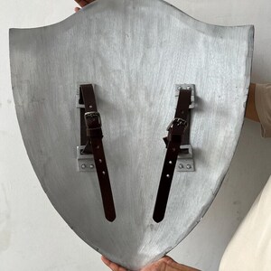 Op de afbeelding: Een zilverkleurig schild met een gebogen bovenkant en twee bruine leren riemen met gespen. Het schild heeft een houten textuur. Het schild wordt door een persoon omhoog gehouden.