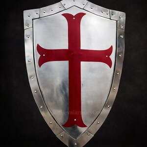 May include: A silver-colored shield with a riveted border and a prominent red cross design. The shield has a pointed bottom and a curved top, with a dark background.