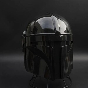 May include: A glossy black Mandalorian helmet, a replica of the iconic helmet from the Star Wars series. The helmet is displayed on a wire stand against a dark background. The helmet's design features a rounded top and angular facial features.
