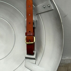 Puede incluir: Un escudo circular de metal plateado con un dise&ntilde;o liso de anillos conc&eacute;ntricos. En el interior, dos soportes de metal plateado fijados con pernos sostienen una correa de cuero marr&oacute;n con una hebilla de metal en tono lat&oacute;n.