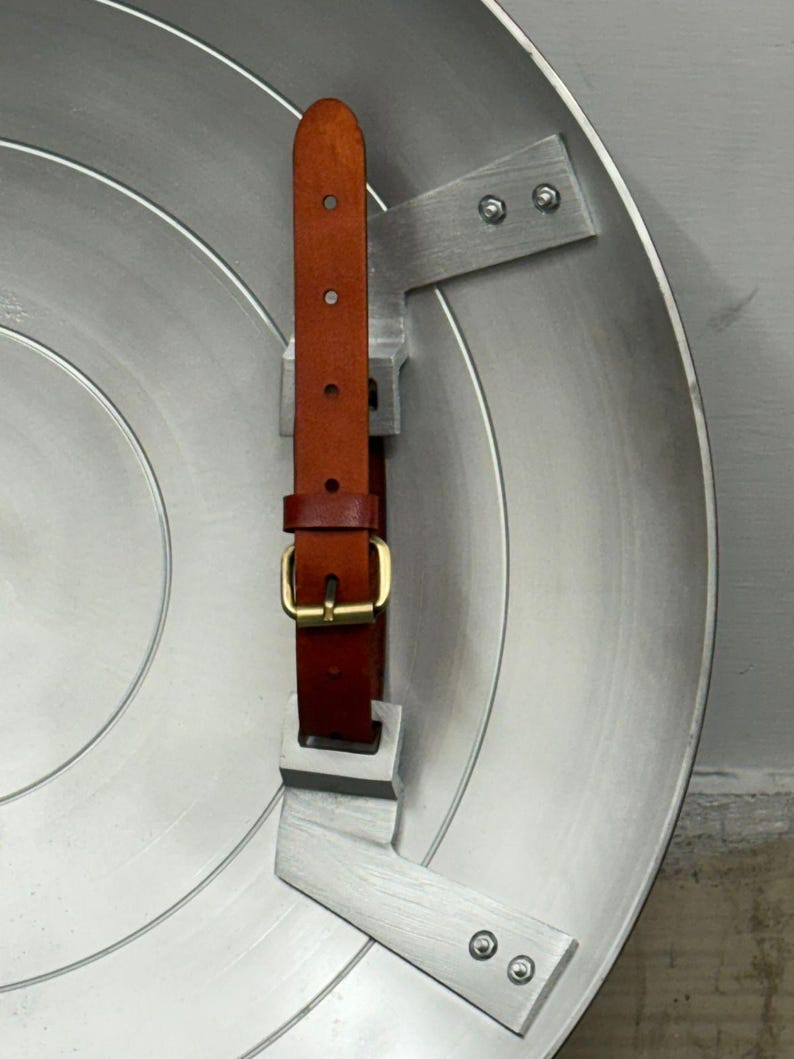 Puede incluir: Un escudo circular de metal plateado con un dise&ntilde;o liso de anillos conc&eacute;ntricos. En el interior, dos soportes de metal plateado fijados con pernos sostienen una correa de cuero marr&oacute;n con una hebilla de metal en tono lat&oacute;n.