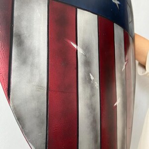 Op de afbeelding: Een schild met een patriottisch ontwerp, met rode en witte strepen en een blauw bovenste gedeelte met een zilveren ster. Het schild heeft een licht versleten uiterlijk, met zichtbare krassen en slijtage.