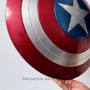 May include: A red, white, and blue shield with a silver star in the center. The shield has a metallic finish and a circular design. The shield is held by a hand. The text "MEDIEVAL ARMOR" is visible.