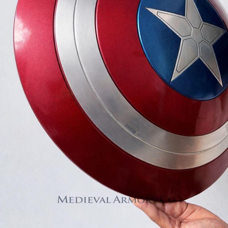May include: A red, white, and blue shield with a silver star in the center. The shield has a metallic finish and a circular design. The shield is held by a hand. The text "MEDIEVAL ARMOR" is visible.