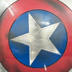 Puede incluir: Un escudo circular con una estrella plateada central sobre un fondo azul vibrante. El dise&ntilde;o incluye un anillo rojo ancho que rodea el centro azul, bordeado por un borde exterior plateado. La superficie tiene un acabado met&aacute;lico desgastado.