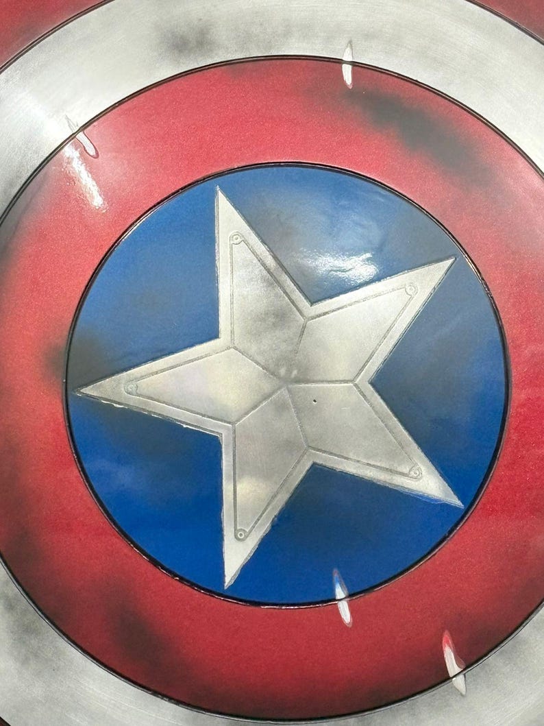 Puede incluir: Un escudo circular con una estrella plateada central sobre un fondo azul vibrante. El dise&ntilde;o incluye un anillo rojo ancho que rodea el centro azul, bordeado por un borde exterior plateado. La superficie tiene un acabado met&aacute;lico desgastado.
