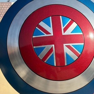 Op de afbeelding: Een rond schild met een blauwe buitenring, een zilveren middenring en een rood centrum. Het centrum heeft een blauw en wit Union Jack-ontwerp. Het schild is een replica van een iconisch accessoire van een superheld.