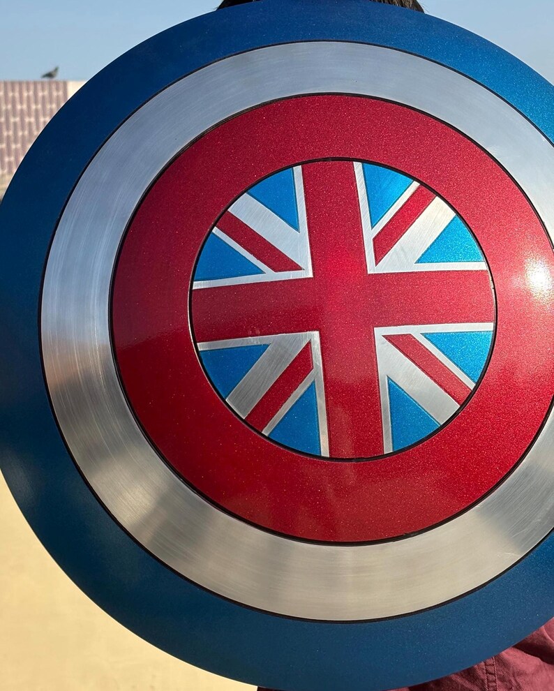 Op de afbeelding: Een rond schild met een blauwe buitenring, een zilveren middenring en een rood centrum. Het centrum heeft een blauw en wit Union Jack-ontwerp. Het schild is een replica van een iconisch accessoire van een superheld.