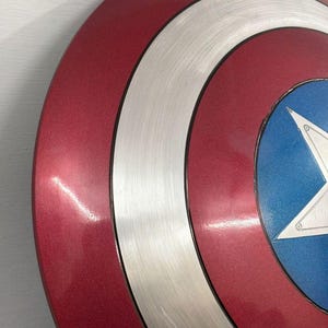 May include: A circular shield featuring concentric rings of metallic red and brushed silver, with a central blue circle containing a large white five-pointed star. The surface has a smooth, reflective finish with visible circular graining on the silver sections.