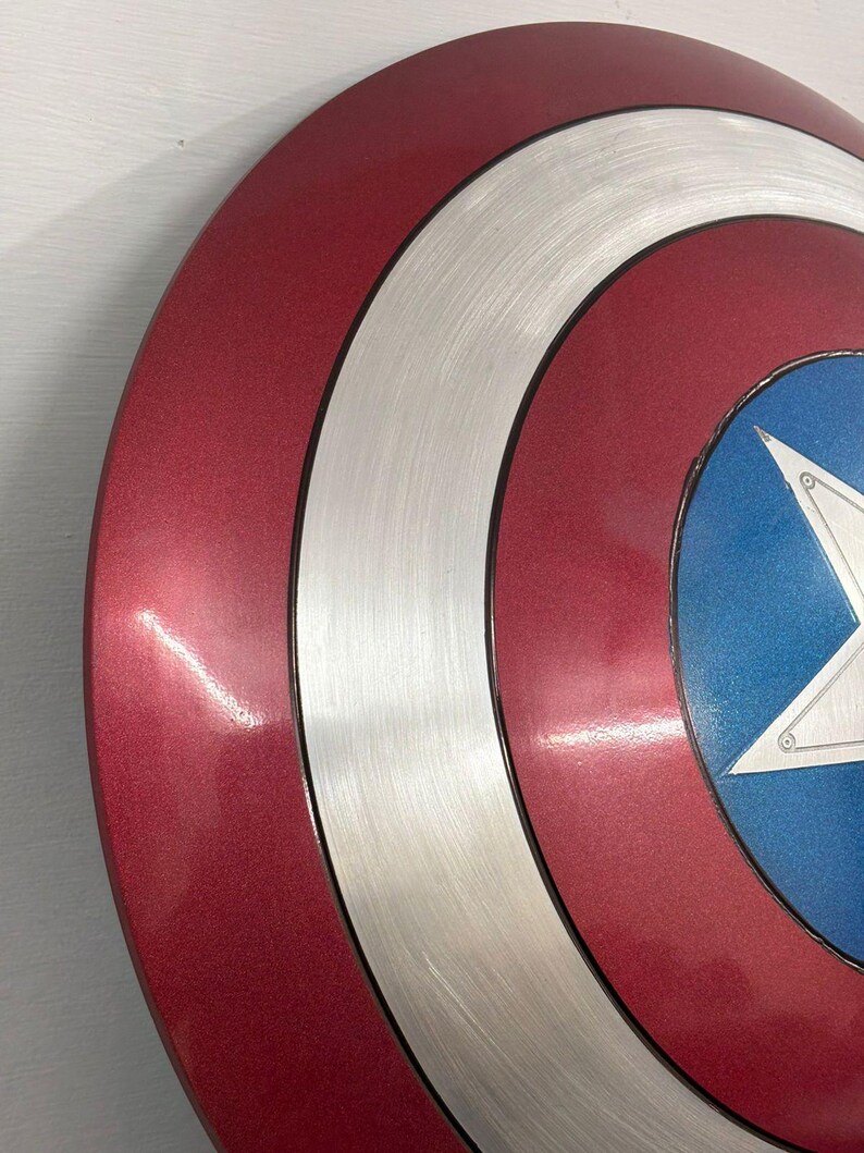 May include: A circular shield featuring concentric rings of metallic red and brushed silver, with a central blue circle containing a large white five-pointed star. The surface has a smooth, reflective finish with visible circular graining on the silver sections.