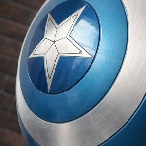 Op de afbeelding: Een close-up van een Captain America-schild. Het schild is voornamelijk blauw met een zilveren ster in het midden. Zilveren banden omcirkelen de ster en de buitenrand van het schild. Het schild heeft een metallic glans.