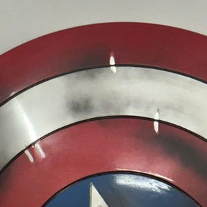 Puede incluir: Un escudo circular con anillos conc&eacute;ntricos rojos y plateados, con un c&iacute;rculo azul central que contiene una estrella plateada. La superficie tiene un acabado desgastado y envejecido con manchas oscuras y reflejos de luz sobre el metal.