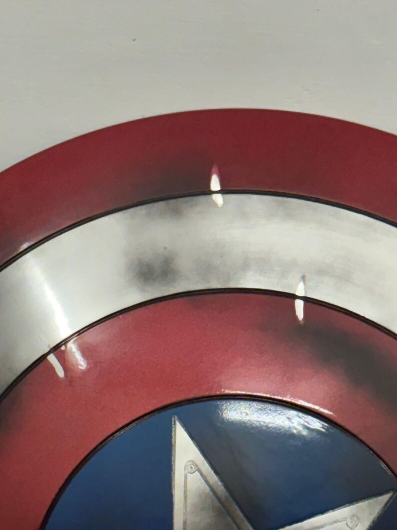 Puede incluir: Un escudo circular con anillos conc&eacute;ntricos rojos y plateados, con un c&iacute;rculo azul central que contiene una estrella plateada. La superficie tiene un acabado desgastado y envejecido con manchas oscuras y reflejos de luz sobre el metal.