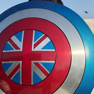 Op de afbeelding: Een cirkelvormig schild met een Union Jack-ontwerp. Het schild heeft een blauwe buitenring, een zilveren middenring en een rode kern. De Union Jack is afgebeeld in blauw, wit en rood. Het schild heeft een metallic afwerking.