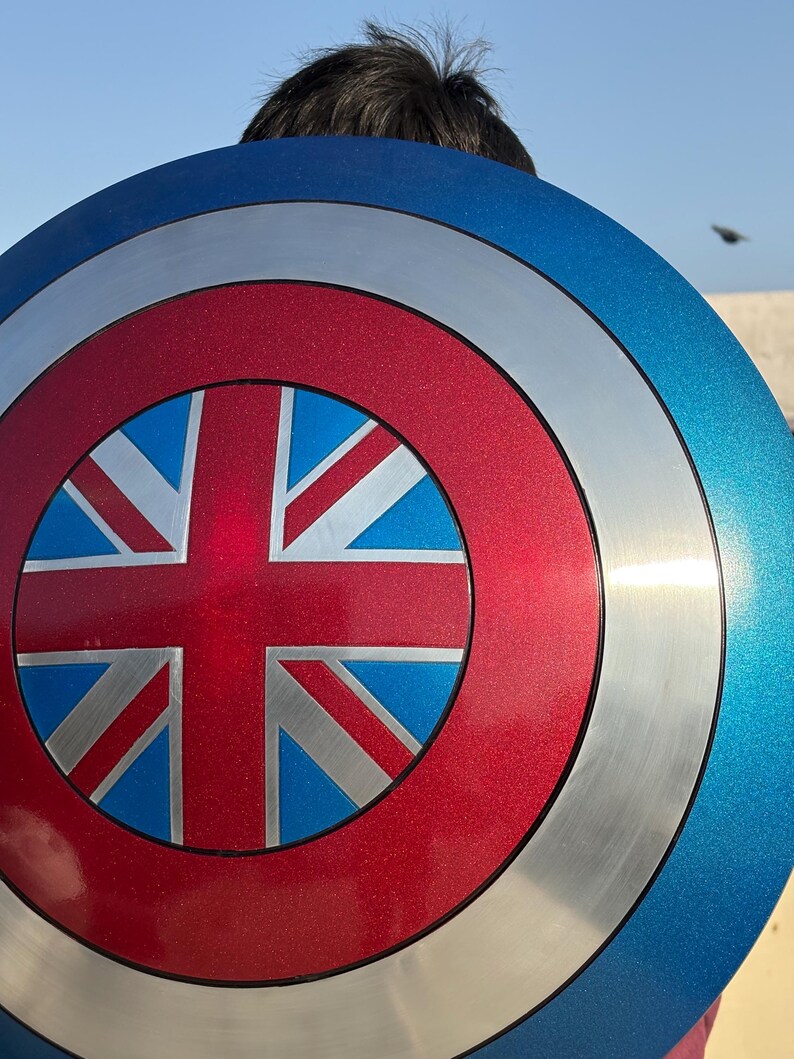 Op de afbeelding: Een cirkelvormig schild met een Union Jack-ontwerp. Het schild heeft een blauwe buitenring, een zilveren middenring en een rode kern. De Union Jack is afgebeeld in blauw, wit en rood. Het schild heeft een metallic afwerking.