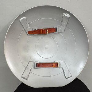 May include: A circular silver-colored shield featuring a smooth, metallic finish. The interior side displays two silver-toned metal brackets, each fitted with a brown leather strap and a brass-colored metal buckle for securing the shield to an arm.