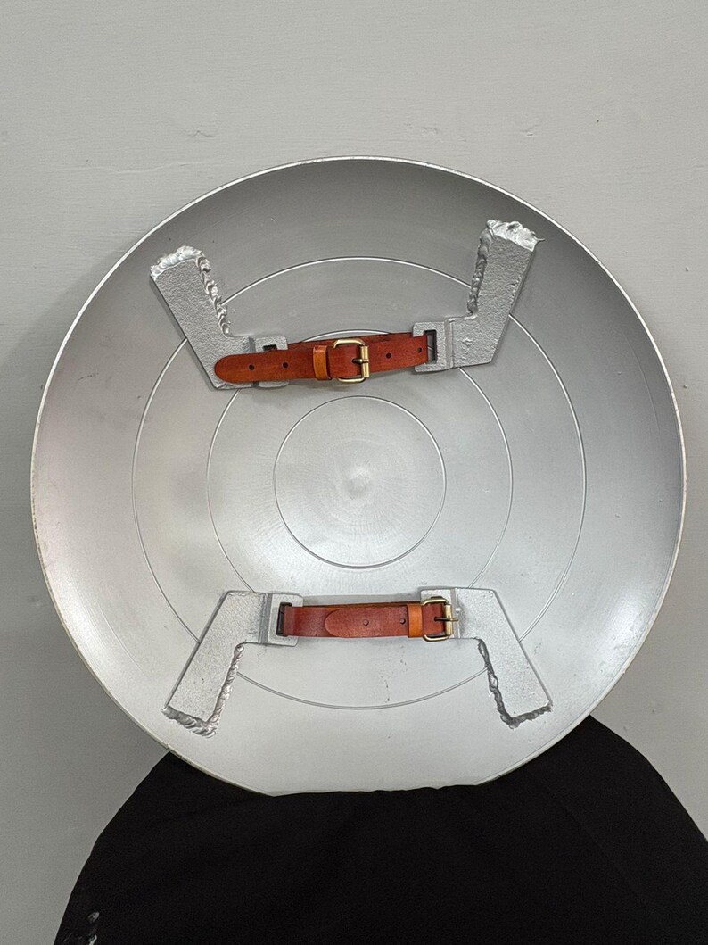 May include: A circular silver-colored shield featuring a smooth, metallic finish. The interior side displays two silver-toned metal brackets, each fitted with a brown leather strap and a brass-colored metal buckle for securing the shield to an arm.