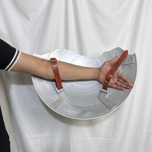 以下が含まれることがあります： 半円形のデザインの銀色の盾。腕に固定するためのバックル付きの2本の茶色の革ストラップが付いています。盾はテクスチャ加工された表面で、コスチュームやコスプレ用に設計されています。直径は約40cmです。