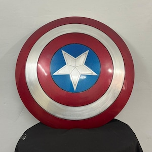 May include: A circular shield featuring a metallic red outer ring, a silver middle ring with a brushed texture, and a central blue circle containing a large, five-pointed silver star. The design is symmetrical with clean, defined color sections.
