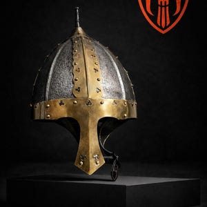 Puede incluir: Un yelmo medieval con acabado plateado y dorado, con una prominente protección facial y remaches decorativos. El yelmo se exhibe sobre un pedestal negro sobre un fondo oscuro. El logotipo de MedievalarmoryCo es visible.