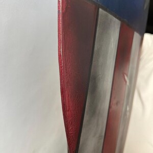 Op de afbeelding: Een close-up van een schild met een rood, wit en blauw kleurenschema. Het schild heeft een verweerde afwerking, met zichtbare slijtage. Het ontwerp omvat rode en witte strepen en een blauw veld met witte sterren.