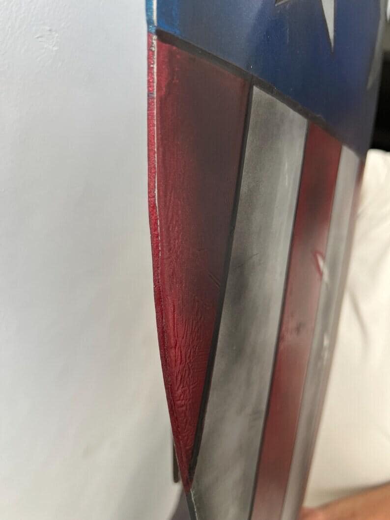 Op de afbeelding: Een close-up van een schild met een rood, wit en blauw kleurenschema. Het schild heeft een verweerde afwerking, met zichtbare slijtage. Het ontwerp omvat rode en witte strepen en een blauw veld met witte sterren.