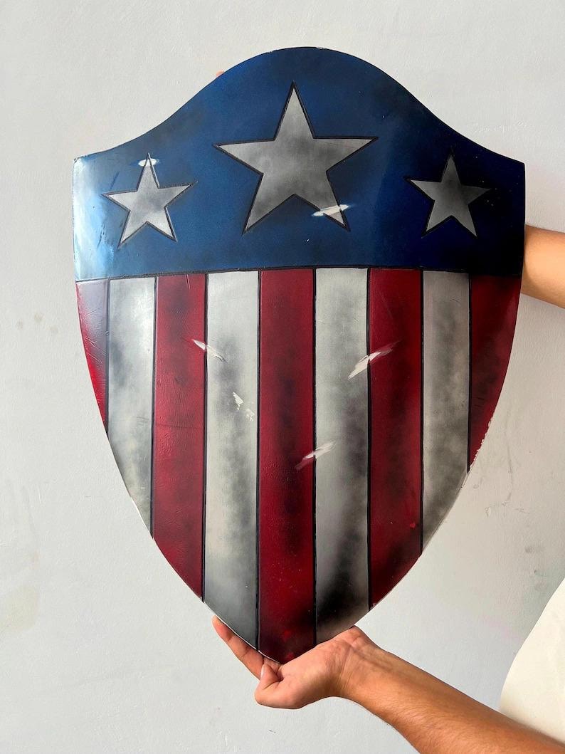 Op de afbeelding: Een schild met een blauw bovenstuk met drie witte sterren. Het schild heeft rode en witte verticale strepen op het onderste gedeelte. Het schild wordt door een persoon vastgehouden.