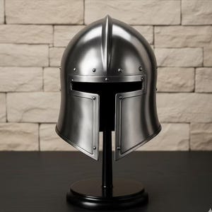 May include: A silver-colored medieval helmet with a visor, rivets, and a rounded top. The helmet is displayed on a black stand with a circular base, set against a light-colored brick wall.