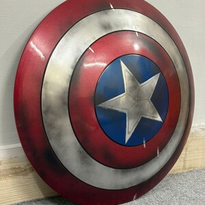 Puede incluir: Un escudo circular con una estrella plateada central sobre un fondo azul, rodeado por un anillo plateado y un anillo exterior rojo. La superficie tiene un acabado desgastado y envejecido con manchas oscuras y texturas met&aacute;licas visibles.