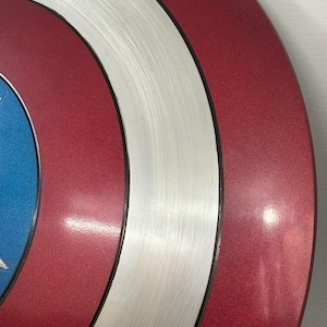May include: A circular shield featuring concentric rings of metallic red and brushed silver, with a vibrant blue center section. The design consists of a smooth, reflective red outer ring, a textured brushed silver middle ring, and a blue inner circle.